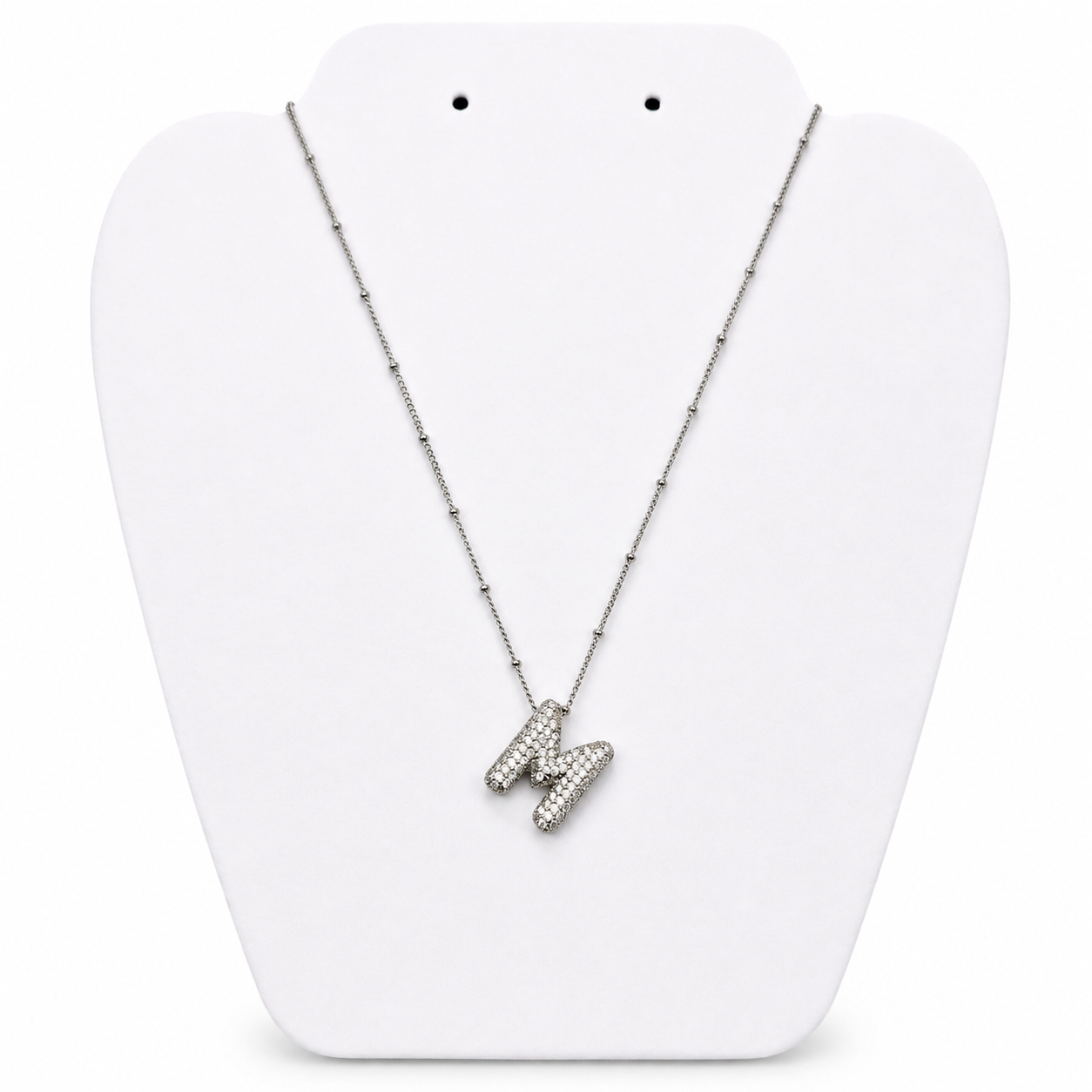 A silver initial necklace featuring the letter 'M' paved with small clear stones, displayed on a white jewelry bust.