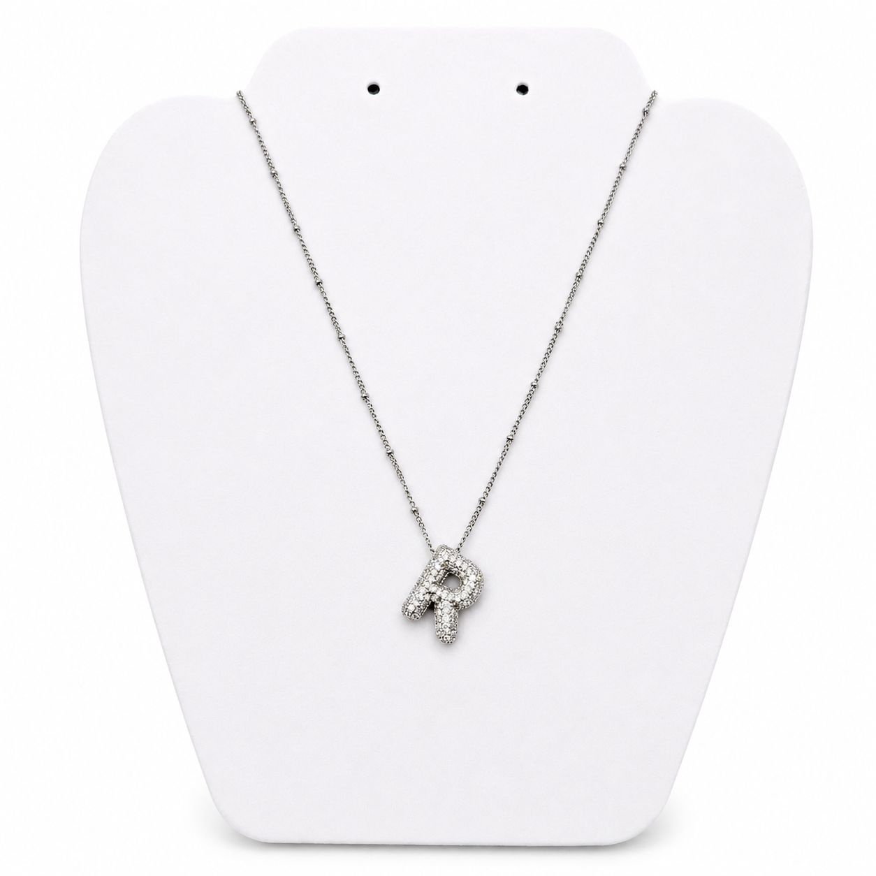 A silver initial necklace featuring the letter 'R' paved with small clear stones, displayed on a white plastic jewelry bust.